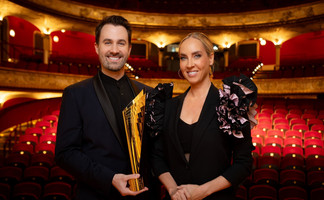 @ ORF / Hans Leitner Ein Mann und eine Frau in eleganter schwarzer Abendgarderobe stehen lächelnd in einem prachtvollen Theater mit rotem Samtinterieur. Der Mann hält eine goldene Trophäe in den Händen.