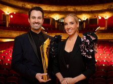 Ein Mann und eine Frau in eleganter schwarzer Abendgarderobe stehen lächelnd in einem prachtvollen Theater mit rotem Samtinterieur. Der Mann hält eine goldene Trophäe in den Händen.