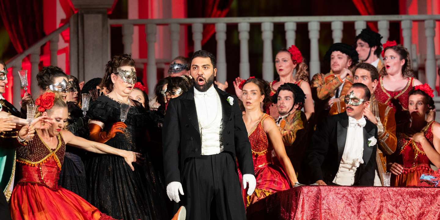 © Anna Stöcher Opernszene von "La Traviata" mit Sänger im Frack und maskierten Darstellerinnen in prunkvollen Kostümen auf Bühne.