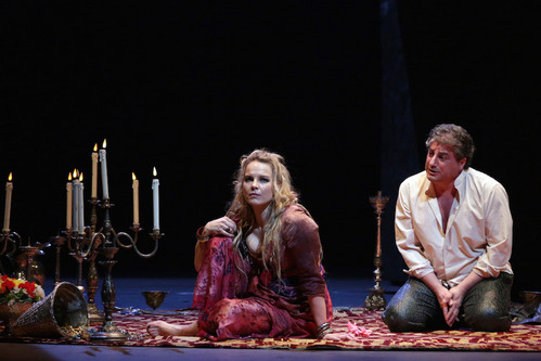 © Marco Brescia & Rudy Amisano Opernszene mit zwei Sänger:innen auf einer Bühne, sitzend auf einem Teppich neben Kerzenleuchtern in dramatischer Lichtstimmung.