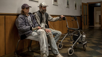  Zwei Menschen mit Kappen sitzen mit Rollator auf Bank in Flur
