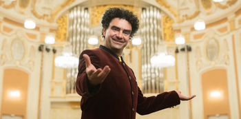 Rolando Villazón steht lächelnd in einem prunkvollen Konzertsaal mit Orgel im Hintergrund und öffnet einladend die Arme in Richtung Publikum.