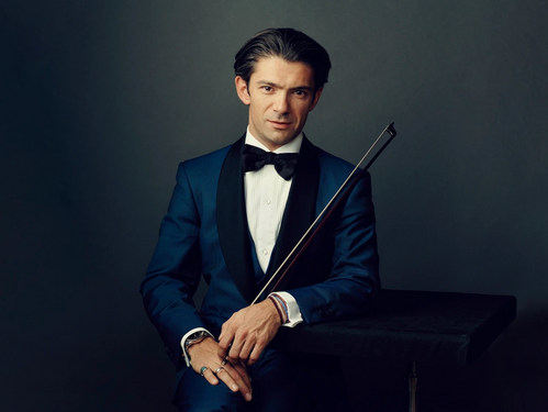 © Anoush Abrar Eleganter Mann im dunkelblauen Smoking mit schwarzer Fliege, der einen Cellobogen hält, vor neutralem Hintergrund.