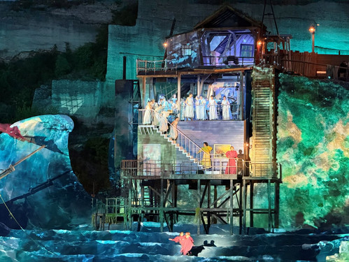 © Adam Kürth, klassikticket.at Abendliche Szene auf einer imposanten Freilichtbühne mit projizierten Wasser- und Wellenmotiven, auf der ein mehrstöckiges Haus mit Treppe und Darsteller:innen in hellen Kostümen zu sehen ist.