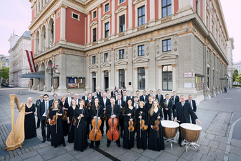 Ein Orchester in eleganter schwarzer Kleidung steht mit Instrumenten vor dem Wiener Musikverein. Im Hintergrund ist die reich verzierte Fassade des historischen Gebäudes mit rot-beiger Farbe und österreichischen Fahnen zu sehen.