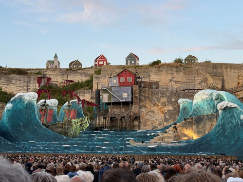 © Adam Kürth, klassikticket.at Tageslichtaufnahme einer spektakulären Bühnenlandschaft mit Wellenattrappen und Piratenschiff vor einer Felskulisse, im Vordergrund ein dichtes Publikum.