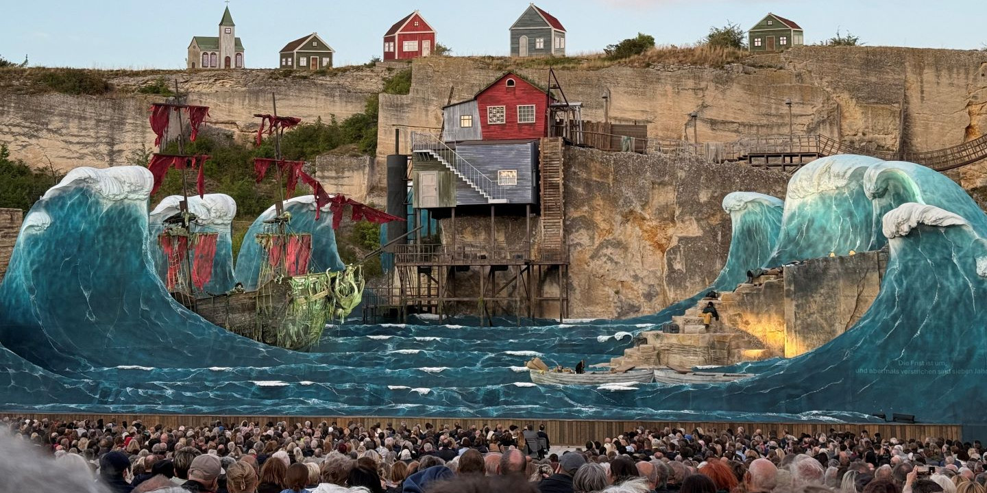 © Adam Kürth, klassikticket.at Tageslichtaufnahme einer spektakulären Bühnenlandschaft mit Wellenattrappen und Piratenschiff vor einer Felskulisse, im Vordergrund ein dichtes Publikum.