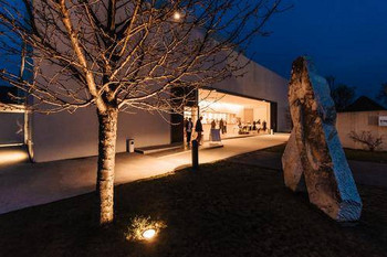 © Rudi Dellinger Abendliche Außenaufnahme eines beleuchteten Veranstaltungsgebäudes mit offenen Glastüren. Gäste treffen ein, im Vordergrund steht ein beleuchteter Baum.