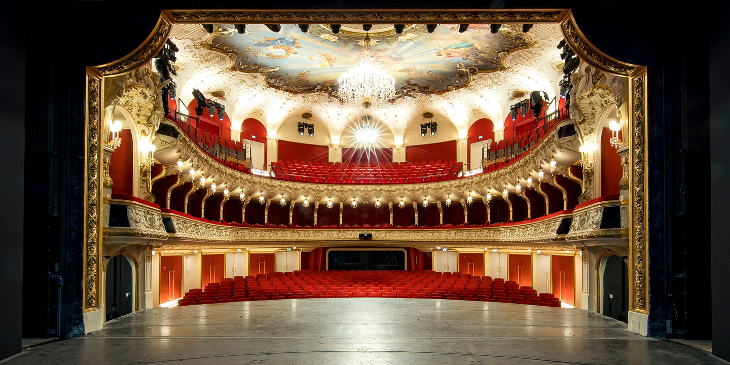 Blick von der Bühne in den prachtvollen Zuschauerraum eines Theaters mit rotem Samt, goldenen Verzierungen, Kronleuchter und Deckenmalerei. Die Sitzreihen sind leer, die Architektur wirkt historisch und opulent.