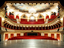 Blick von der Bühne in den prachtvollen Zuschauerraum eines Theaters mit rotem Samt, goldenen Verzierungen, Kronleuchter und Deckenmalerei. Die Sitzreihen sind leer, die Architektur wirkt historisch und opulent.