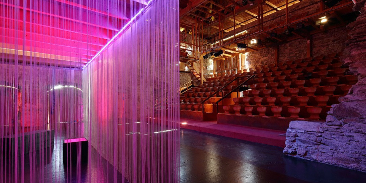 Moderne Theatertribüne mit roten Sitzreihen in rustikaler Stein- und Holzkulisse, im Vordergrund ein mit pink-violettem Licht beleuchteter Vorhang aus Fäden.