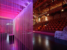 Moderne Theatertribüne mit roten Sitzreihen in rustikaler Stein- und Holzkulisse, im Vordergrund ein mit pink-violettem Licht beleuchteter Vorhang aus Fäden.