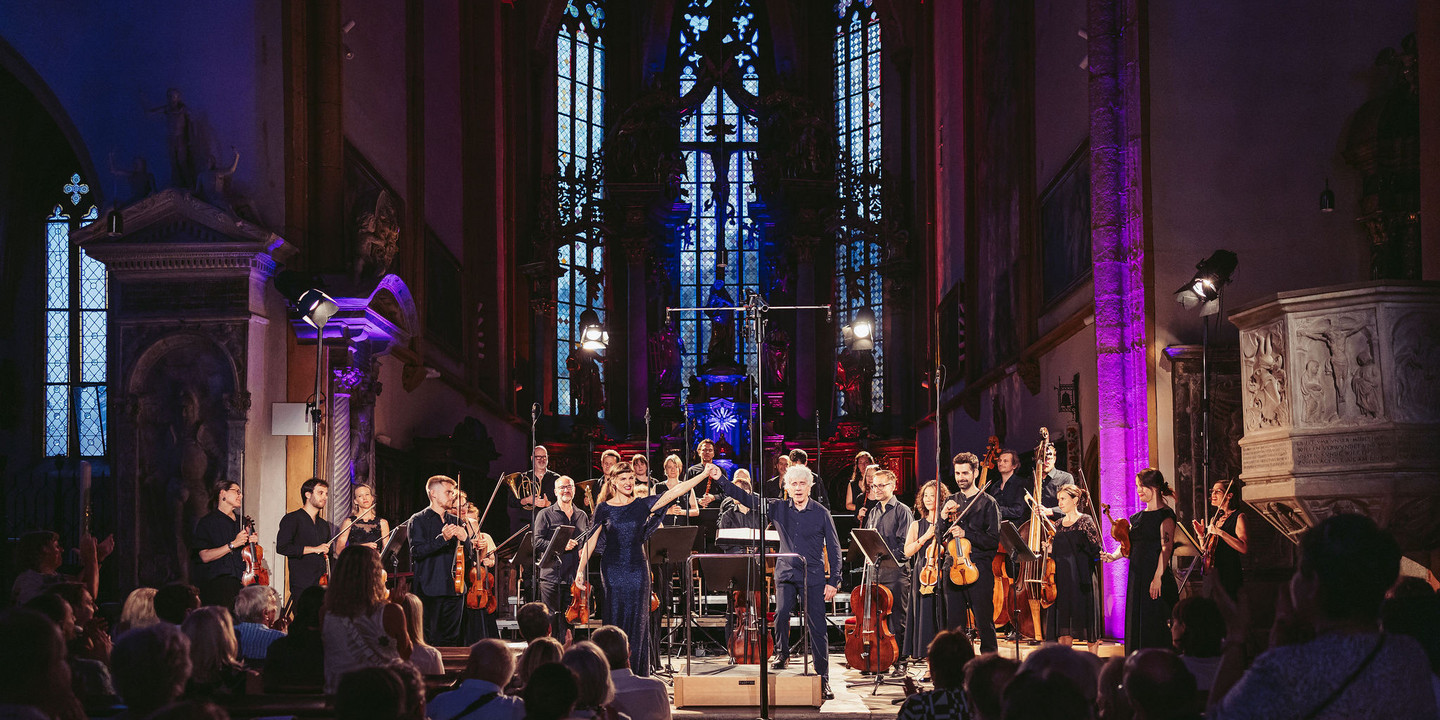 © Marta Gillner Konzertszene in einer prunkvollen Kirche mit hochaufragenden Fenstern und gotischem Altarraum: Ein Orchester mit Streicher:innen, Bläser:innen und Dirigent verbeugt sich gemeinsam mit einer Sängerin im blauen Abendkleid vor dem applaudierenden Publikum. Bühne und Raum sind in farbiges Licht getaucht.