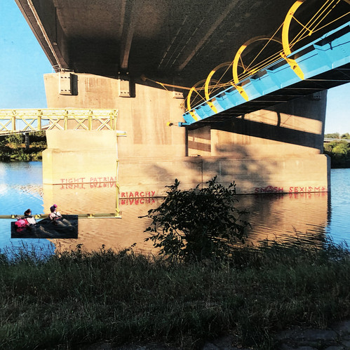Unter einer Brücke mit gelbem Geländer paddeln zwei Menschen in einem Boot über den ruhigen Fluss