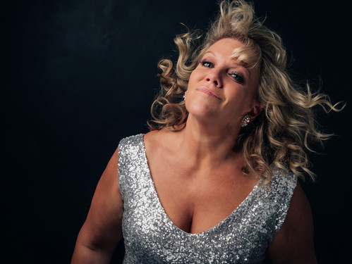 © Becky Lee Frau mit lockigem blondem Haar in glitzerndem silbernen Kleid posiert vor dunklem Hintergrund.